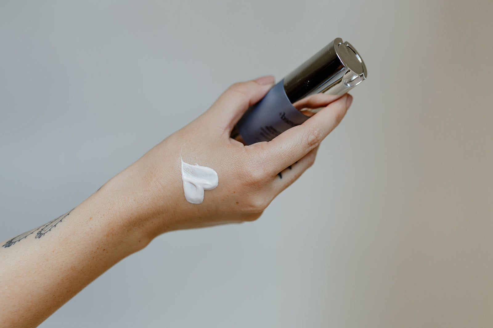 Hand holding a bottle of skincare product with a small amount on the wrist against a neutral background